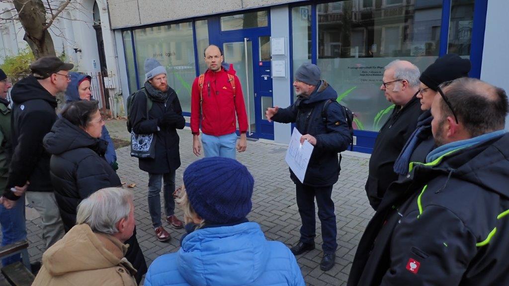 Eine Gruppe Menschen steht im Kreis beieinander. Ein Mann erklärt etwas anhand einer Schautafel.